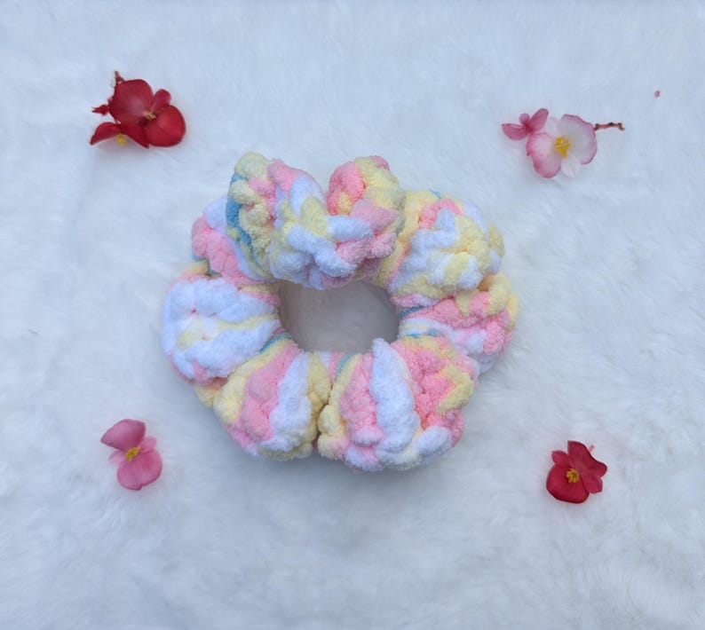May include: A handmade, pastel-colored scrunchie with a soft, textured appearance. The scrunchie features a mix of pink, yellow, blue, and white yarn. The background is a white, fluffy surface, with scattered red and pink flower petals.