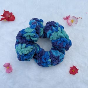 May include: A handmade, plush scrunchie in shades of blue and teal. The scrunchie is made of a soft, textured material and is surrounded by small, colorful flowers.