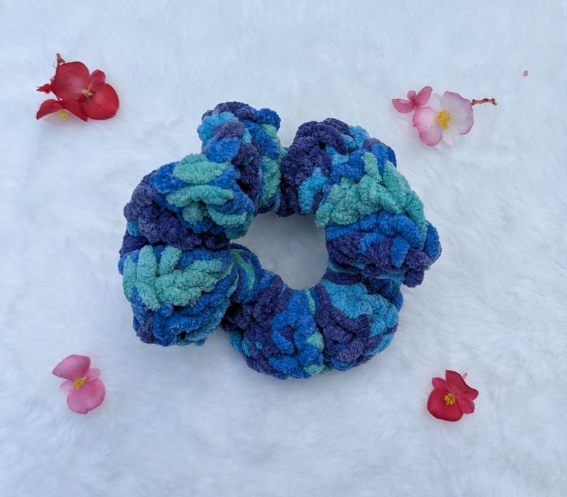 May include: A handmade, plush scrunchie in shades of blue and teal. The scrunchie is made of a soft, textured material and is surrounded by small, colorful flowers.