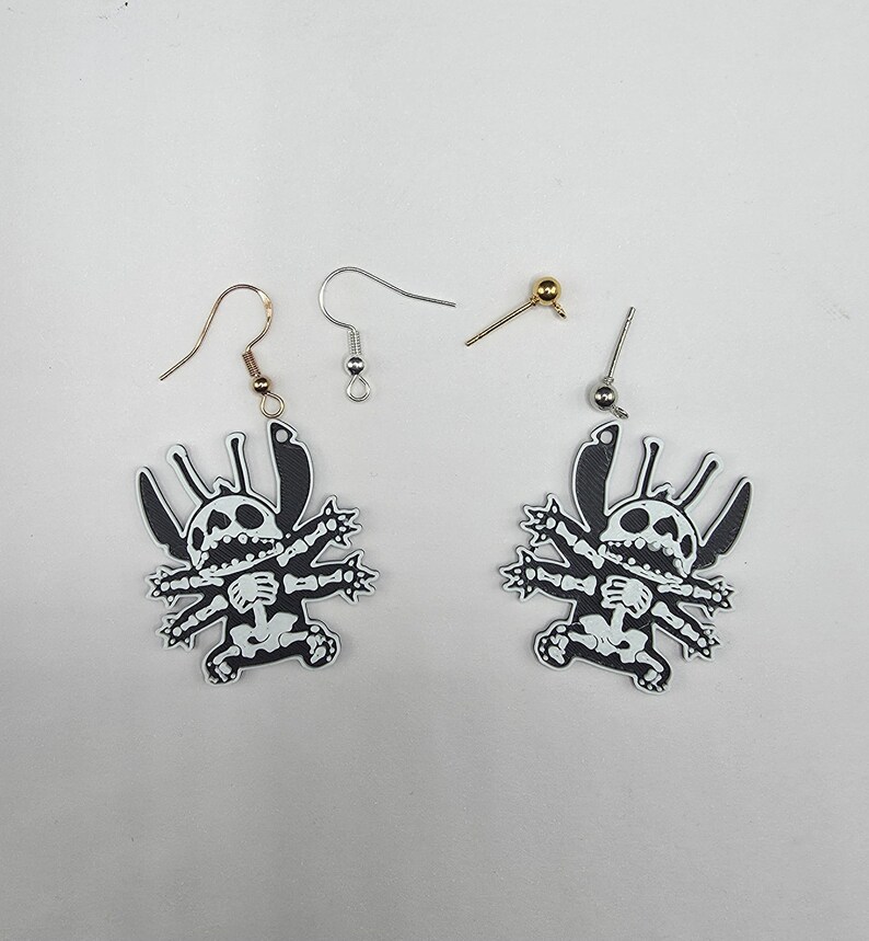 May include: A pair of black and white Stitch-themed earrings with a skeleton design. The earrings have a cartoon-style design and come with various earring hooks and posts. The earrings are displayed on a white surface.