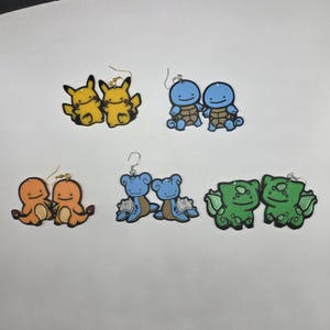 May include: A collection of six pairs of novelty earrings featuring cartoon characters. The earrings include pairs of yellow, blue, orange, and green characters. Each earring has a gold-colored hook.