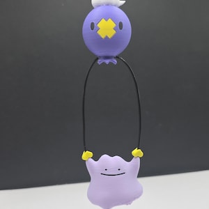 May include: A purple and yellow cartoon character pendant with a black cord. The pendant features a round, purple character with a yellow cross-shaped design and a white cloud-like shape on top. Below, a purple blob with a smiling face is suspended.