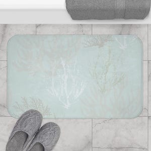 May include: A rectangular bath mat with a light blue background and a coral reef design in shades of white and gray. Gray knitted slippers are at the bottom left. A gray towel is at the top.