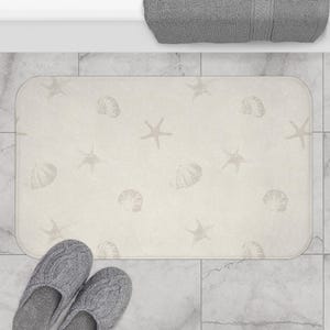 May include: A rectangular beige bath mat with a pattern of seashells and starfish. The mat is placed on a tiled floor, with a pair of gray knitted slippers in the foreground. A gray towel is visible in the background.