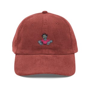 May include: A brick-red corduroy baseball cap featuring an embroidered zombie design. The zombie has gray skin, a pink shirt, and yellow eyes. The cap has a curved brim and an adjustable strap.