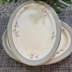 May include: Two oval serving platters with a cream-coloured centre, a grey border, and gold trim. The plates feature delicate grey leaf and berry designs around the edges. The platters are stacked on a textured surface.