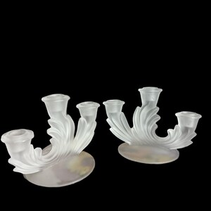 May include: Two frosted glass candle holders. Each holder has three candle cups and a swirling, leaf-like design. The holders sit on oval bases with a subtle, pearlescent finish. The background is black.