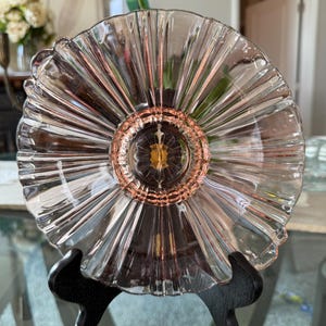 May include: A clear glass plate with a fluted edge and a pink-tinted center. The plate is displayed on a black stand, showcasing its intricate design. The plate's radial pattern and delicate color create an elegant aesthetic.