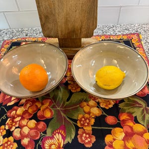 May include: Two beige bowls with silver rims, one containing an orange and the other a lemon, sit on a floral patterned tablecloth. A wooden stand is in the background. The tablecloth features grapes and butterflies.