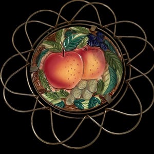 Puede incluir: Un plato decorativo con un diseño de frutas pintadas, que incluye dos manzanas rojas, hojas verdes y uvas. El plato está colocado dentro de un marco de alambre de metal dorado. El diseño de frutas está detallado con varios colores.