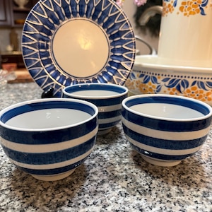 Puede incluir: Colección de vajilla de cerámica azul y blanca. Incluye un plato grande con diseño geométrico azul, tres cuencos pequeños con rayas azules y una jarra con detalles florales naranjas. Sobre una encimera moteada.