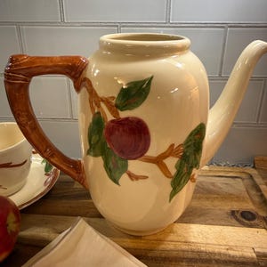 Puede incluir: Tetera de cerámica color crema con asa y boquilla marrones. La tetera está decorada con un diseño de manzana roja y hojas verdes. Se ve una taza y un platillo a juego. La tetera está sobre una superficie de madera.