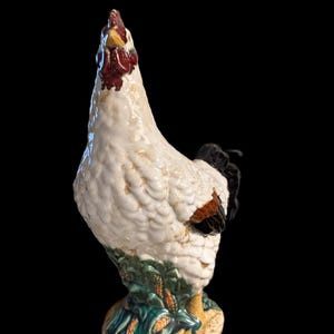 May include: A ceramic rooster figurine with a white body, red comb, and black tail feathers. The base features green leaves and yellow accents. The rooster is posed looking upwards.