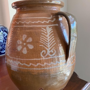 May include: A large, brown ceramic jug with a handle. The jug features white painted floral and geometric designs. The jug is glazed and has a glossy finish. The jug is sitting on a wooden surface.