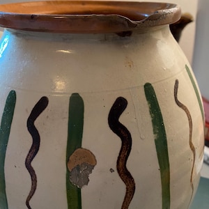 May include: A vintage ceramic pitcher with a cream-colored body, adorned with vertical green and brown wavy stripes. The rim and handle are a warm brown. The pitcher shows signs of age, with some paint loss and wear, adding to its antique charm.