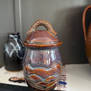 Puede incluir: Jarra de cerámica vidriada con tapa y asa trenzada. La jarra presenta un patrón de ondas y está acabada en tonos marrones, morados y azules. La jarra está sobre un libro con la palabra "TABLE" visible.