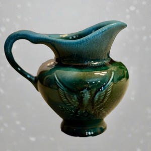 May include: A small, green ceramic pitcher with a handle and a spout. The pitcher has a glossy finish and a decorative embossed design on the front. The handle and rim are a darker shade of green. The pitcher is approximately 6 inches tall.