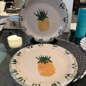 May include: A set of decorative plates with a hand-painted pineapple design. The plates have a cream background with a green and red border. The pineapple is yellow and orange with green leaves. Ideal for kitchen decor.