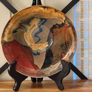 Può includere: Una ciotola in ceramica rotonda caratterizzata da uno smalto multicolore con motivi vorticosi di rosso intenso, blu scuro, marrone terra e crema. La ciotola è esposta su un supporto nero a tre gambe, che ne mette in risalto il bordo ampio.