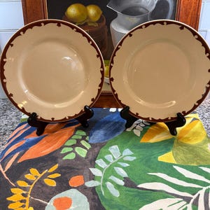 Puede incluir: Dos platos color crema con borde festoneado marrón oscuro se exhiben sobre soportes negros. Los platos descansan sobre un mantel con estampado floral colorido. Un cuadro enmarcado de limones y una jarra está en el fondo.