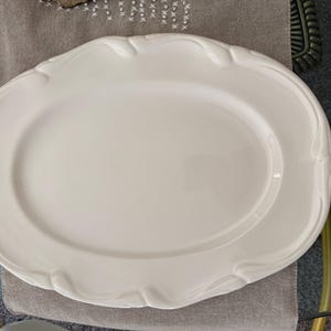 Puede incluir: Una fuente ovalada blanca con borde festoneado. La fuente es de cerámica o porcelana y está colocada sobre una superficie de color neutro. El plato para servir es adecuado para servir alimentos.