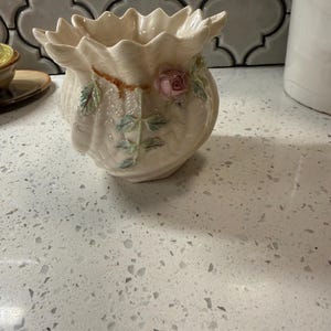 May include: A cream-colored ceramic planter with a scalloped, shell-like rim. The exterior features a raised, three-dimensional design of a small pink rosebud with green leaves and a brown stem, adding texture to the smooth, glossy surface.