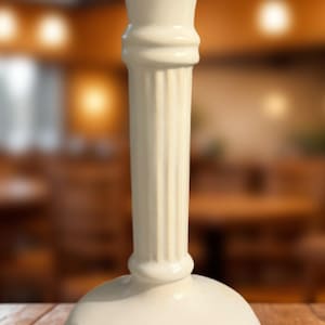 May include: Off-white ceramic candlestick holder with a fluted column design. The base is round and the top flares out. The candlestick holder is a decorative item for home decor.