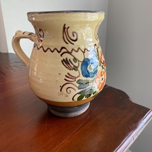 May include: A hand-painted ceramic mug with a cream-colored base and a dark brown rim. The mug features floral designs in blue and orange, with green leaves and brown accents. It has a curved handle and sits on a dark brown base.