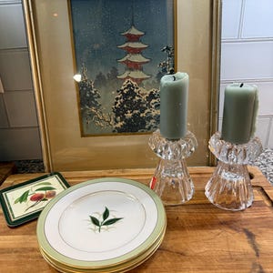 Puede incluir: Una impresión enmarcada de una pagoda japonesa en un paisaje nevado, con dos velas verdes en candelabros de cristal, una pila de platos blancos con borde verde y diseño de hojas, y un posavasos con una ilustración de frutas.