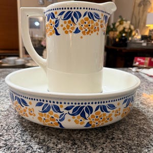 May include: A white ceramic pitcher and basin set. The pitcher has a handle and spout, and the basin is round. Both pieces are decorated with blue and orange floral patterns and blue dots.