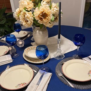 May include: A beautifully set dining table with a navy blue tablecloth. The table is adorned with cream-colored plates with a floral design, blue wine glasses, and a vase of flowers. A black candle sits in a crystal candlestick.