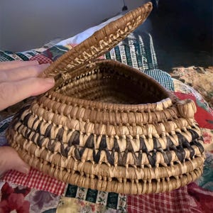 May include: Handmade woven basket with a hinged lid. The basket has a natural, light brown colour with a decorative pattern of dark brown and black woven elements. The lid is partially open, revealing the interior.