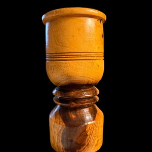 May include: A two-toned wooden pepper mill with a light brown top and a darker brown base. The top section has horizontal grooves, and the base has a rounded shape. The mill is set against a black background.
