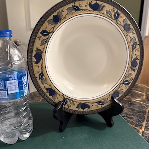 Könnte beinhalten: Eine weiße Keramikschale mit einem dekorativen Rand mit blauen Blumen- und Blättermustern. Die Schale wird auf einem schwarzen Ständer präsentiert, mit einer Wasserflasche im Hintergrund.