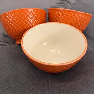 May include: Three orange ceramic bowls with a diamond pattern on the exterior. The interior of the bowls is white. The bowls are arranged on a gray surface.