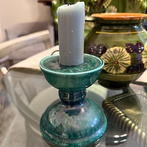 May include: A teal ceramic candle holder with a gray candle. The candle holder has a round base and a flared top. The background includes green ceramic plates and a decorative vase.