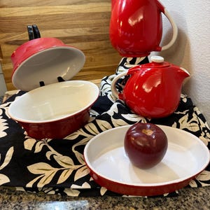 Pode incluir: Uma coleção de utensílios de cozinha vermelhos e brancos, incluindo um jarro, uma chaleira e assadeiras. Uma maçã vermelha está em um prato branco e vermelho. Os itens estão dispostos em um tecido com estampa floral em preto e branco.