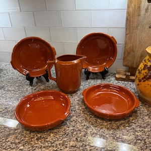 Puede incluir: Un conjunto de utensilios de cocina de cerámica de color terracota, que incluye cuatro platos redondos con asas y una jarra. Los platos son de diferentes tamaños y están sobre soportes negros. La jarra tiene un asa y un pico. Sobre una encimera moteada.