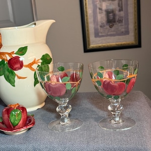 May include: Clear wine glasses with a stem and a round base, decorated with red apples and green leaves. A matching pitcher and a small apple-shaped dish are also visible, all with apple designs. The items are arranged on a gray surface.