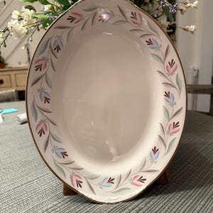May include: An oval ceramic serving platter with a white center and a gold-trimmed rim. The border features a repeating pattern of stylized pink and light blue tulip-like flowers connected by thin, curved grey stems and green leaves.