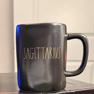 Puede incluir: Una taza de cerámica negra con un asa curvada. La palabra "SAGITTARIUS" está impresa en letras doradas en la parte delantera de la taza. La taza está sobre una superficie oscura.