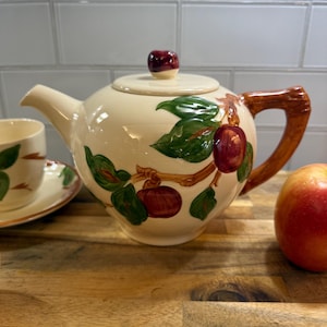 May include: Cream-colored ceramic teapot with a lid and a brown handle. The teapot is decorated with green leaves and red apples. A matching teacup and saucer are visible to the left. An apple is on the right.