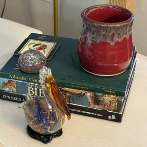 May include: A glass pelican figurine with blue and brown hues sits on a stack of books. A red and brown ceramic mug and a decorative paperweight are also present. The books have green covers and gold lettering.