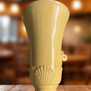 May include: A yellow ceramic vase with a flared top and a pedestal base. The vase features shell and scroll designs. The vase is approximately 10 inches tall and sits on a wooden surface.