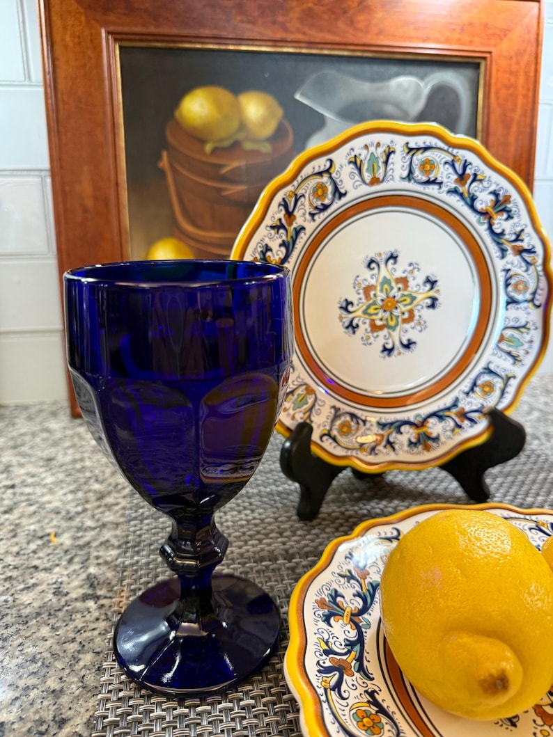 Op de afbeelding: Een kobaltblauwe glazen kelk staat voor twee decoratieve borden met een bloemenontwerp en citroenen. De borden hebben een wit midden, een oranje rand en gele geschulpte randen. Op de achtergrond hangt een schilderij van citroenen in een houten emmer.