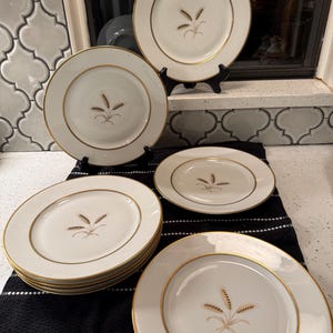 May include: A set of white ceramic plates featuring a gold rim and a central wheat stalk illustration. The collection includes several stacked plates and two upright plates displayed on black stands, all sharing the same wheat motif.