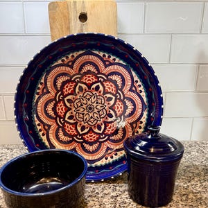 May include: A collection of kitchenware including a large blue decorative plate with a floral design, a small black bowl, and a blue lidded jar. A wooden cutting board is in the background.