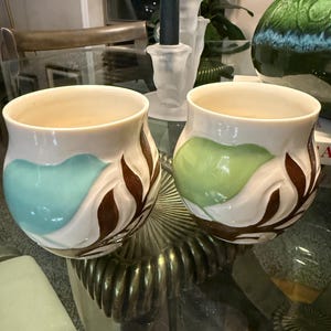 May include: Two ceramic vases with a cream base, featuring blue and green leaf designs and brown accents. The vases have a rounded shape and are displayed on a glass table, showcasing a decorative home decor theme.
