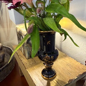 May include: A dark blue ceramic vase with gold trim and floral accents holds a bouquet of pink and purple flowers with green leaves. The vase sits on a distressed wooden surface.
