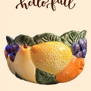 May include: A ceramic fruit bowl with a pear, lemon, and orange design, accented with green leaves and purple grapes. The words "hello fall" are written in a cursive font above the bowl. The bowl is set against a cream background.